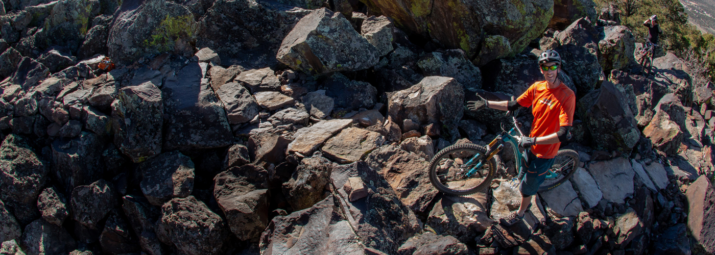 IMBA Trail Solutions staff member Joey Klein smiling while standing over a bike and gesturing to technical rock features on a trail in Cedar City, Utah