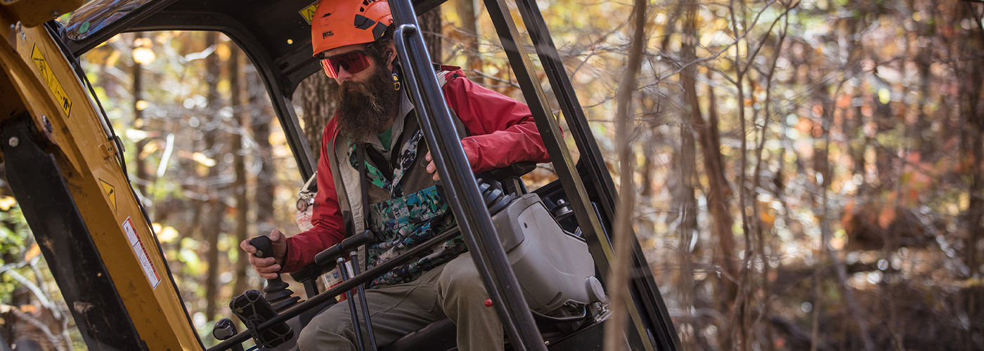 IMBA Trail Solutions staff member operating a mini excavator in a wooded landscape