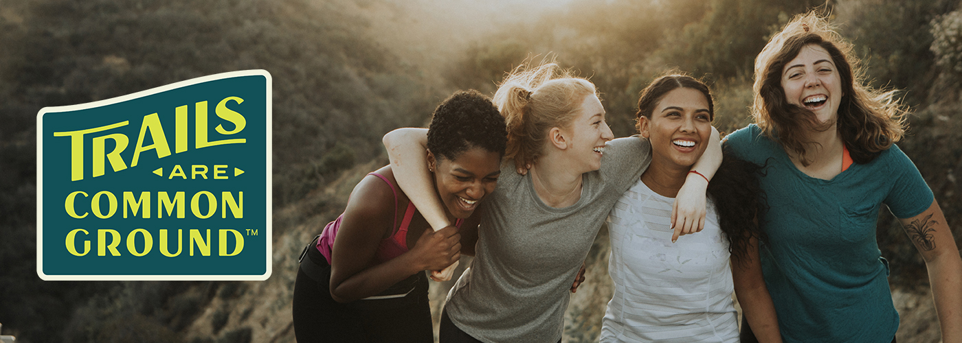 Four female hikers with arms around one another's shoulders smile and laugh on the trail