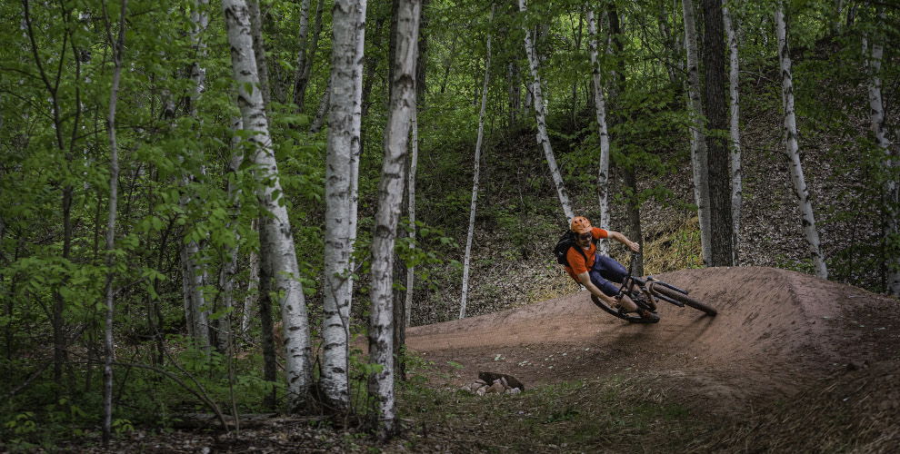 man in red shirt rides bike around a big berm in a birch forest