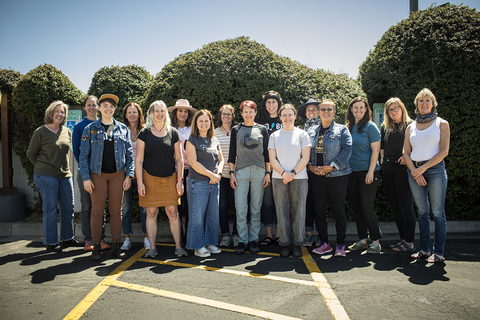 Rad women of the IMBA staff community standing in group photo stance in front of green bushes.