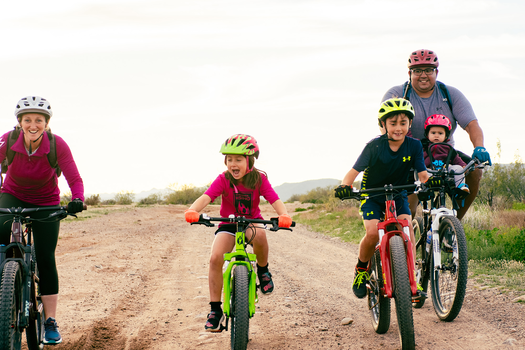 Family ride consisting of an adult female, and adult male riding with a baby, and two young mountain bikers on a gravel road. 