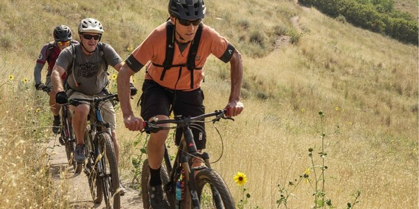 IMBA's Dave Wiens riding on the Bonneville Shoreline Trails with two mountain bikers behind him. Greenish-brown brush on edges of dirt single track. 
