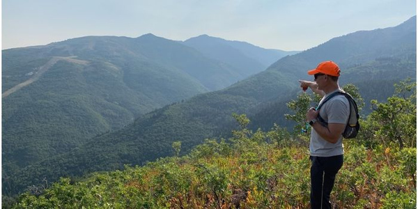 Bob Larsen, a white, middle aged man in a red hat, pointing off into the distance in Utah at green hued mountains. 